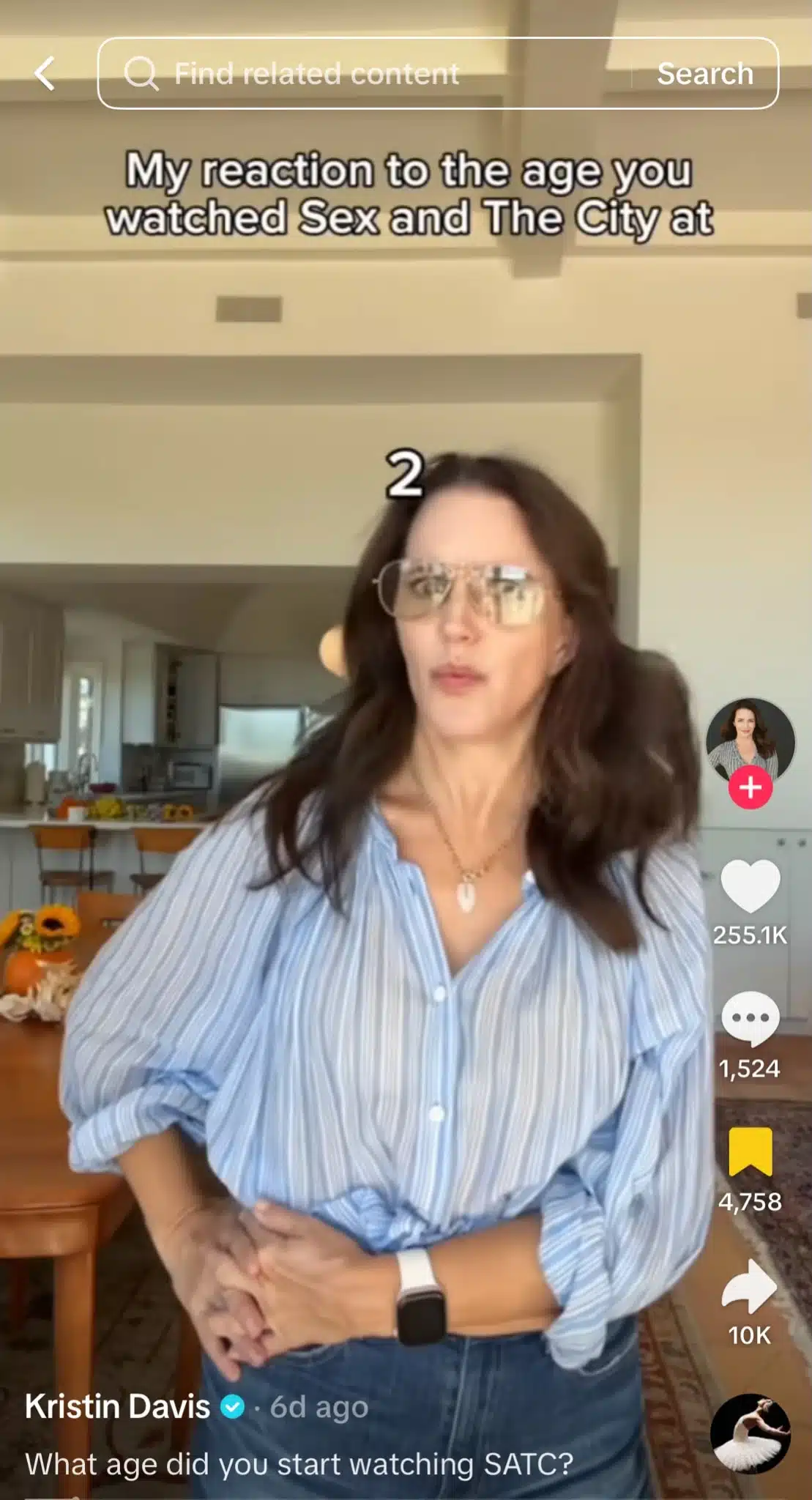 TikTok video from Kristin Davis showing her wearing glasses and a blue striped shirt while making a surprised expression in her kitchen. Text on screen reads “My reaction to the age you watched Sex and The City at,” with the number 2 above her head.